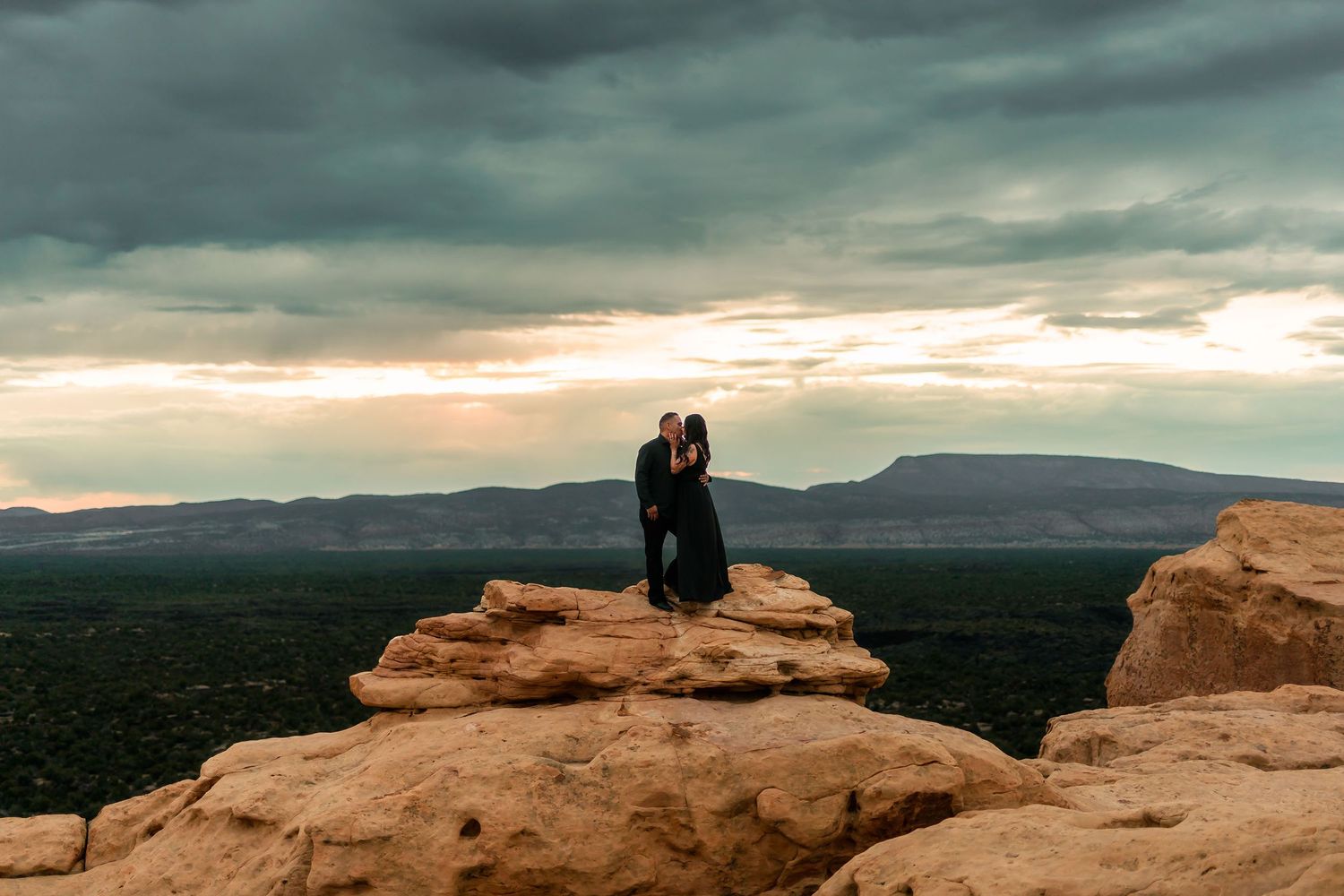 Couple in black embracing on desert rock formation under moody dramatic cloudy sunset sky.