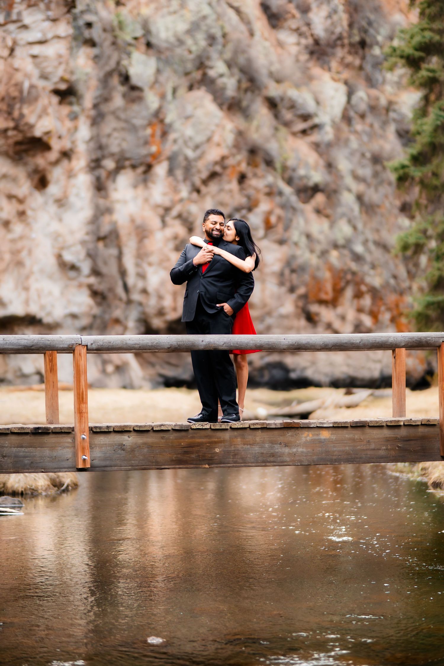 A couple shares a romantic embrace while standing on a wooden bridge over water with rocky cliffs behind them.