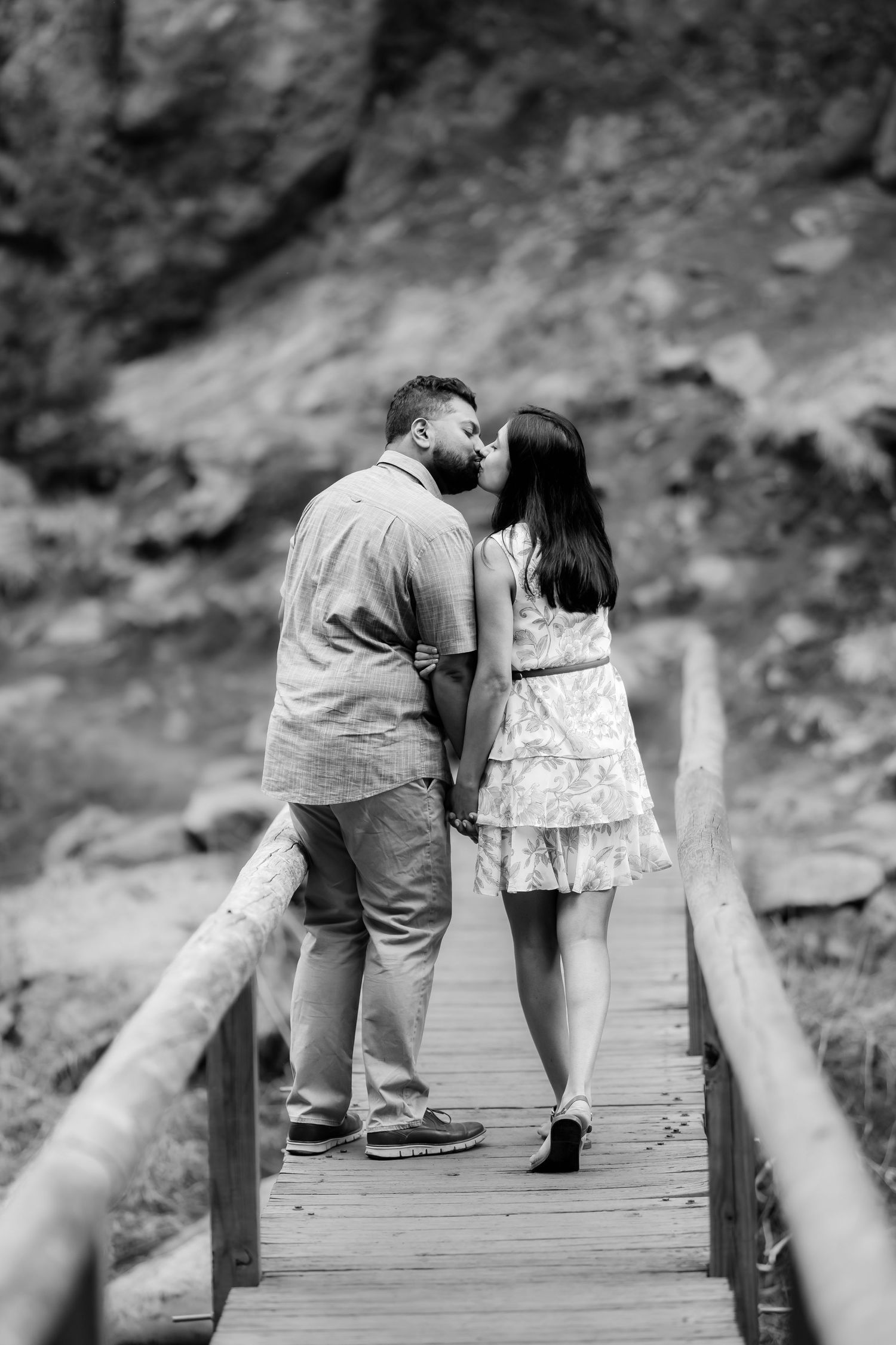A couple shares an intimate moment while standing on a wooden bridge surrounded by rocky cliffs in monochrome.