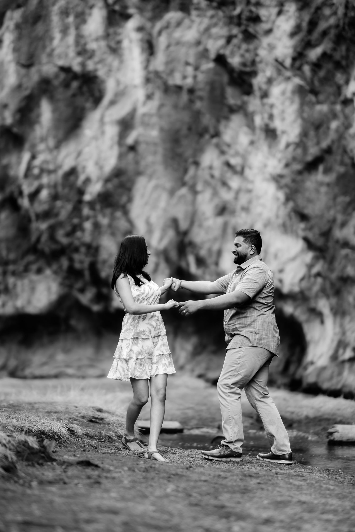A couple dancing playfully by rocky cliffs near a stream in black and white.