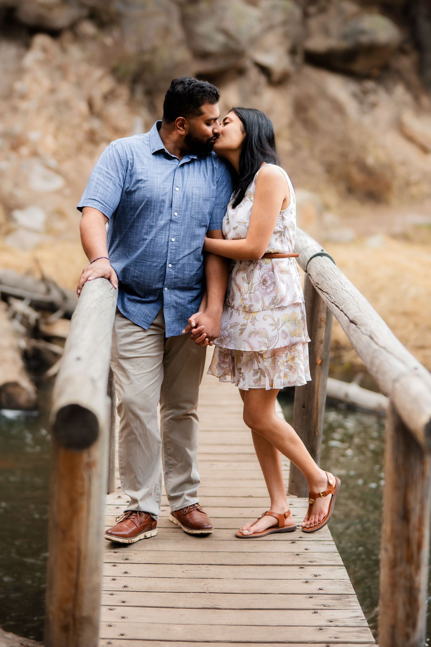 A couple shares an intimate moment on a wooden footbridge over a creek in a natural setting.