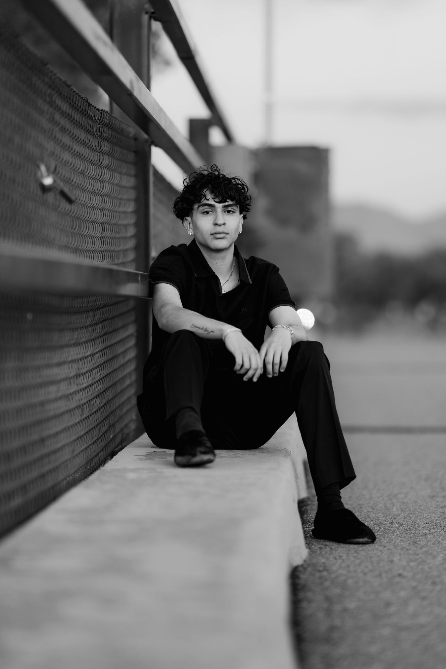 A black and white photo of someone sitting by a chain link fence wearing dark clothes in an urban setting.