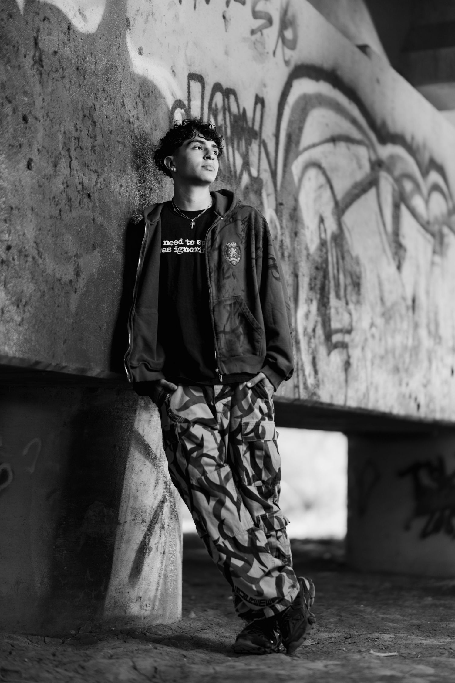 Black and white portrait of someone in camo pants and jacket leaning against a graffiti covered concrete wall.