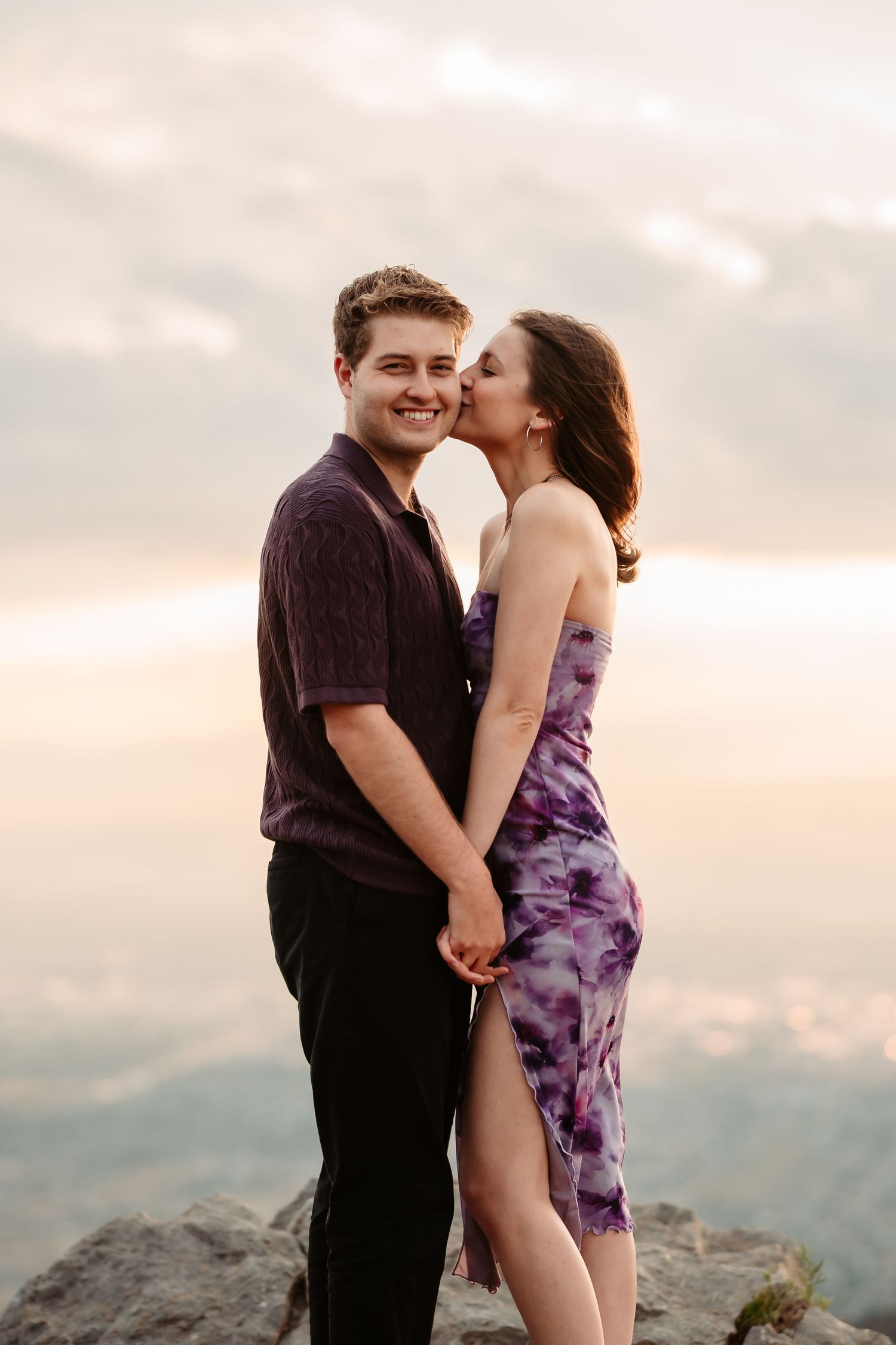 A romantic couple embraces against a soft, cloudy sunset backdrop on a scenic mountaintop overlook.