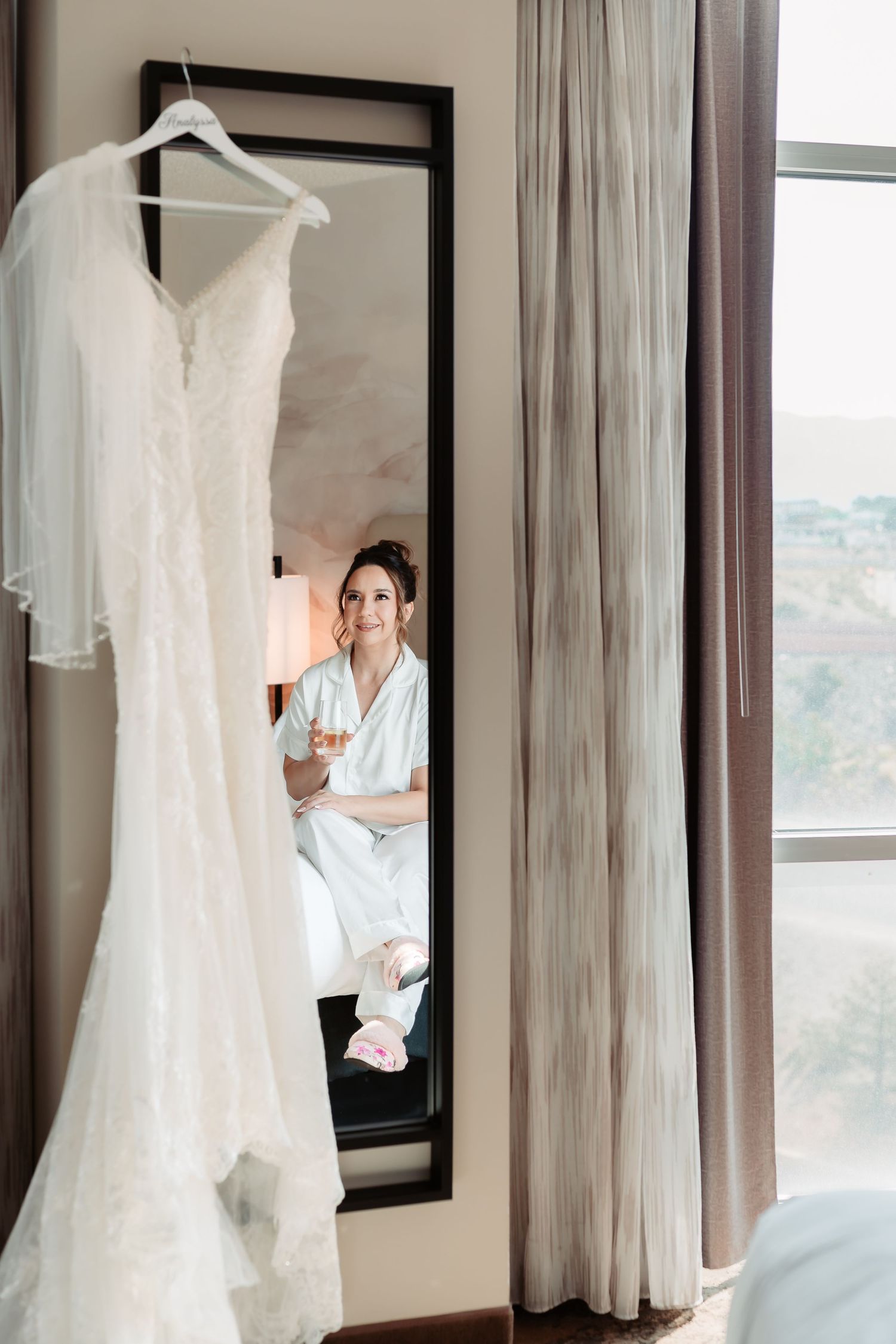 White lace wedding dress hanging on display in a room with natural light and sheer curtains.