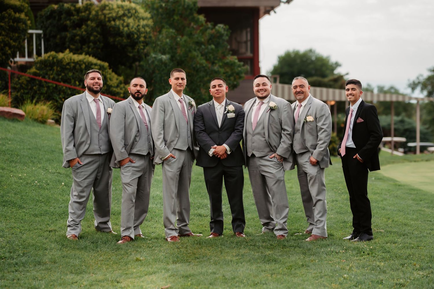 Wedding party in gray suits poses casually outdoors on lush grass during summer celebration.