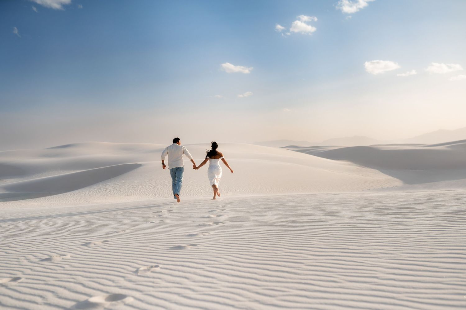 Two people walk hand in hand across pristine white sand dunes under a soft cloudy sky at sunset.