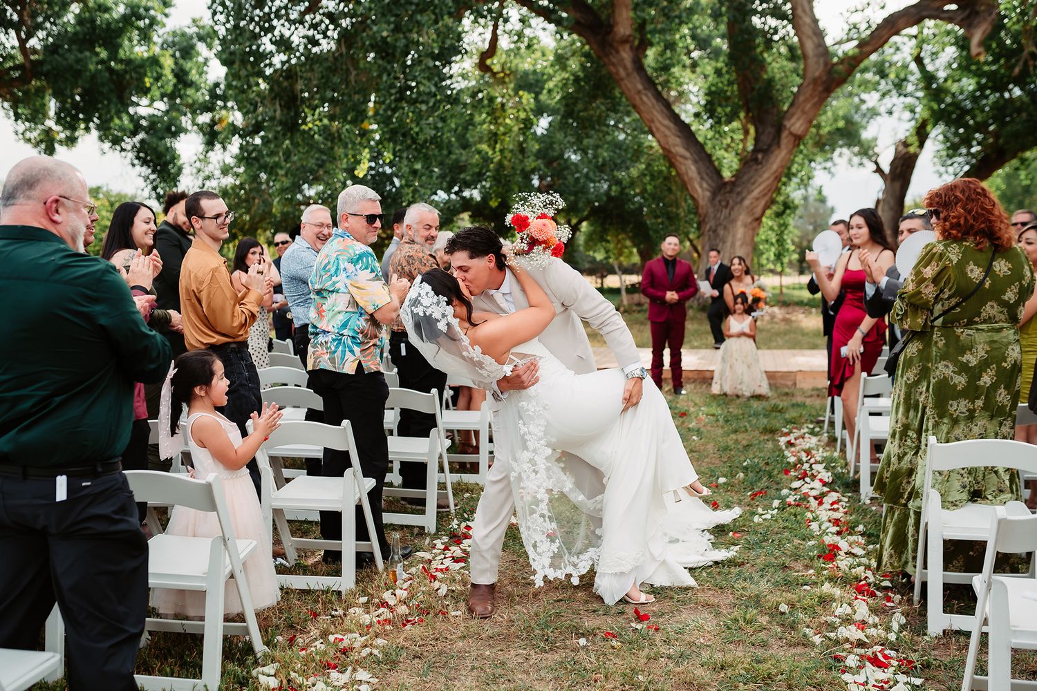 A joyful outdoor wedding ceremony under trees with white chairs and scattered rose petals on the ground.
