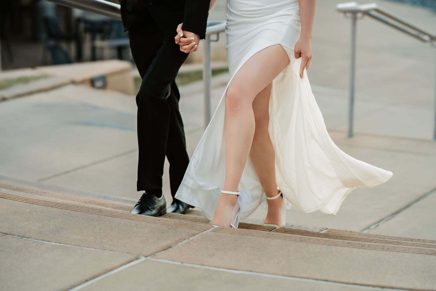 A couple in wedding attire walks together on a city sidewalk with the bride's dress flowing in the breeze.