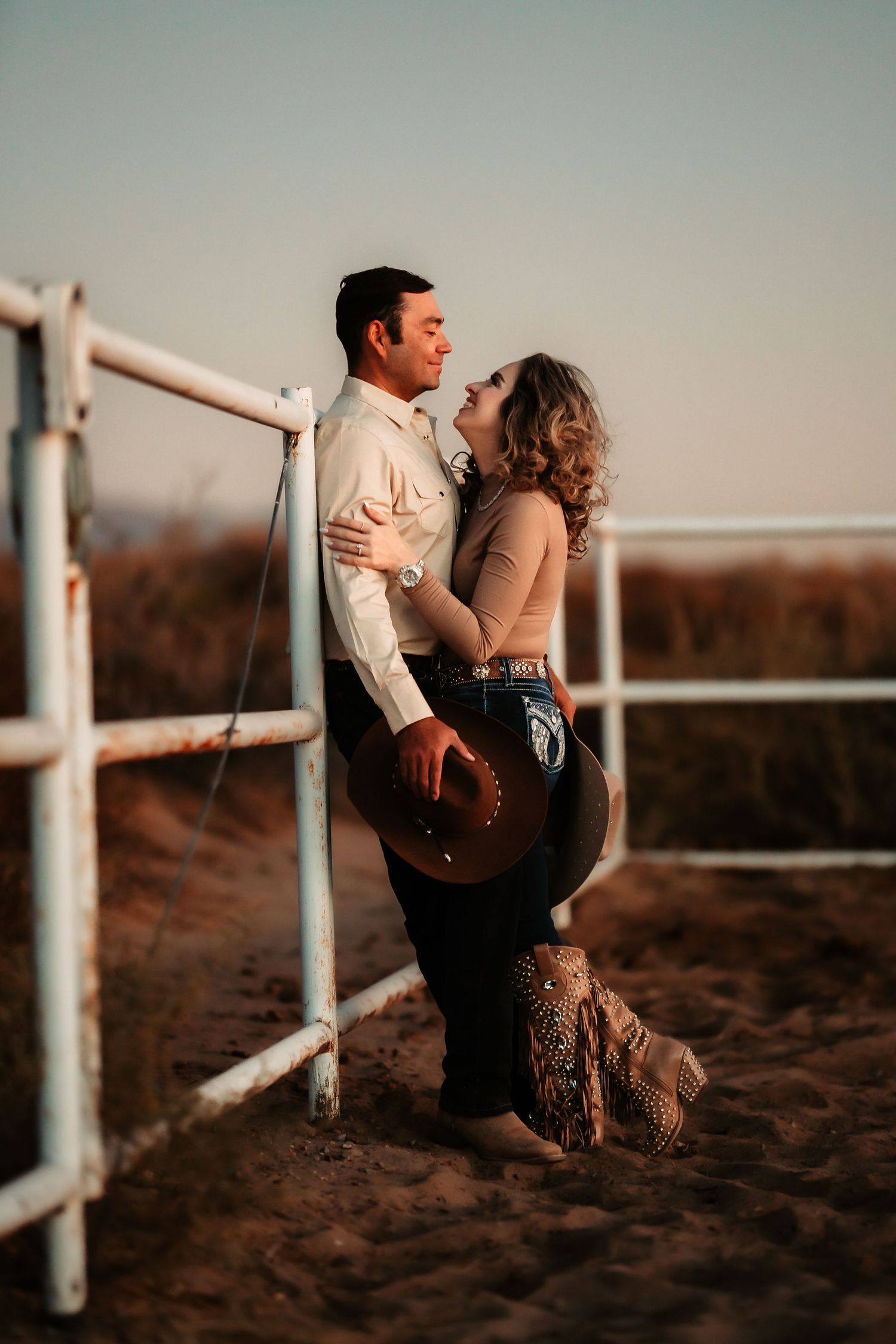 A romantic couple sits together on a white fence at sunset in a rustic countryside setting.