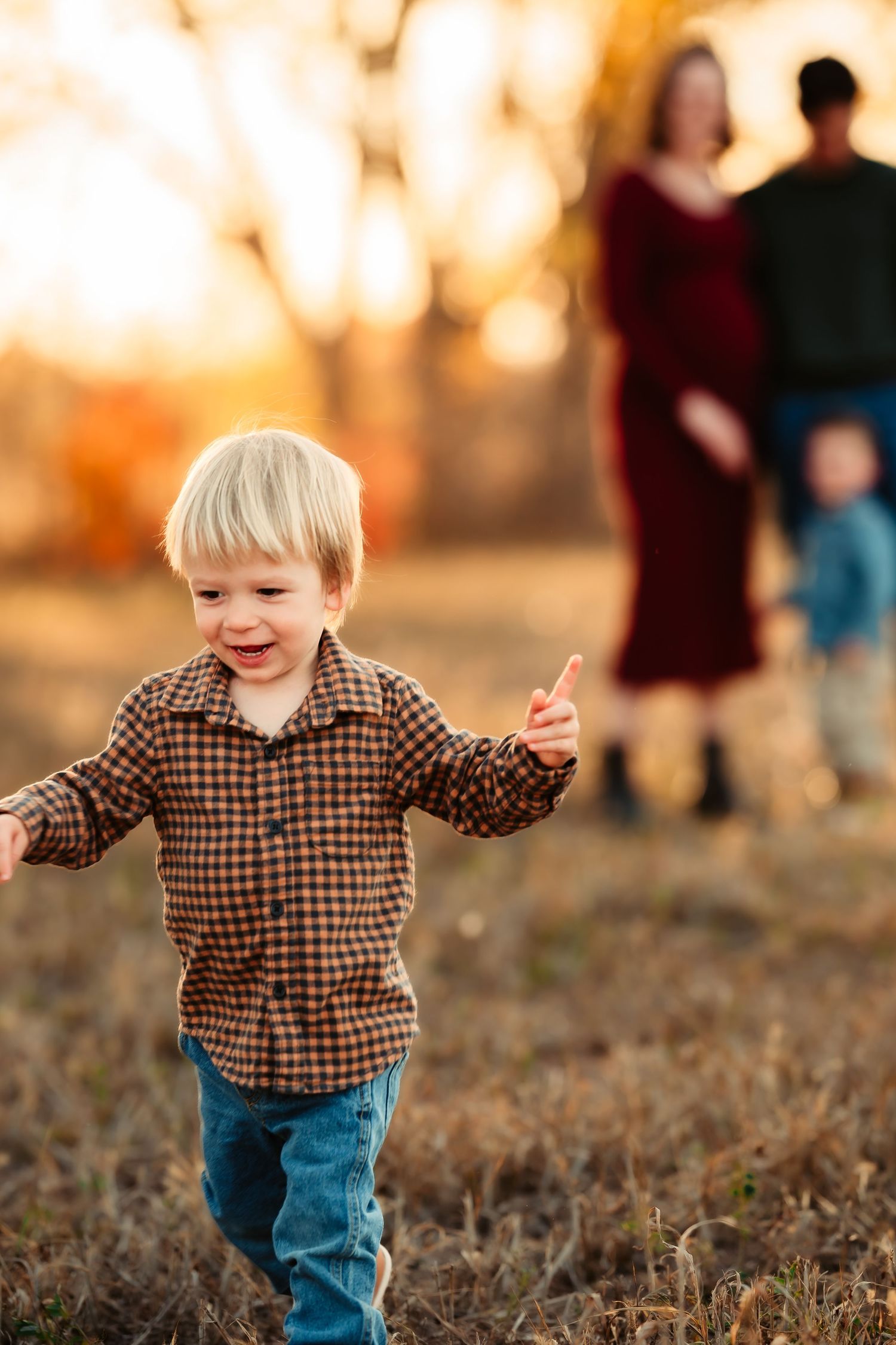 Young child in plaid shirt runs joyfully through a field at sunset while others stand in the background.