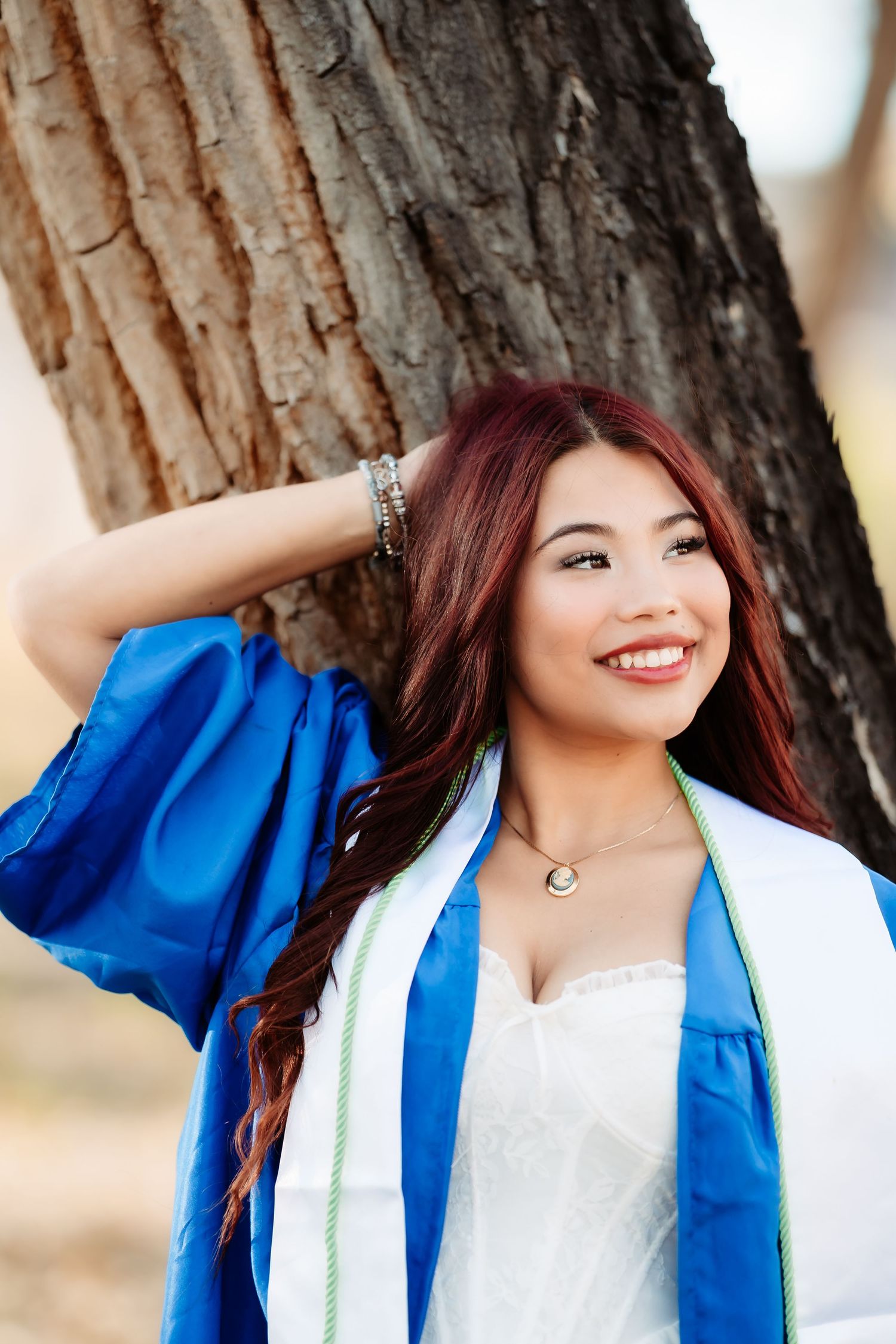 Graduate in blue academic regalia and white stole poses confidently outdoors near tree.