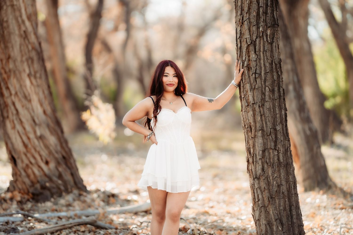 Graduate in white dress poses between trees in natural woodland setting.