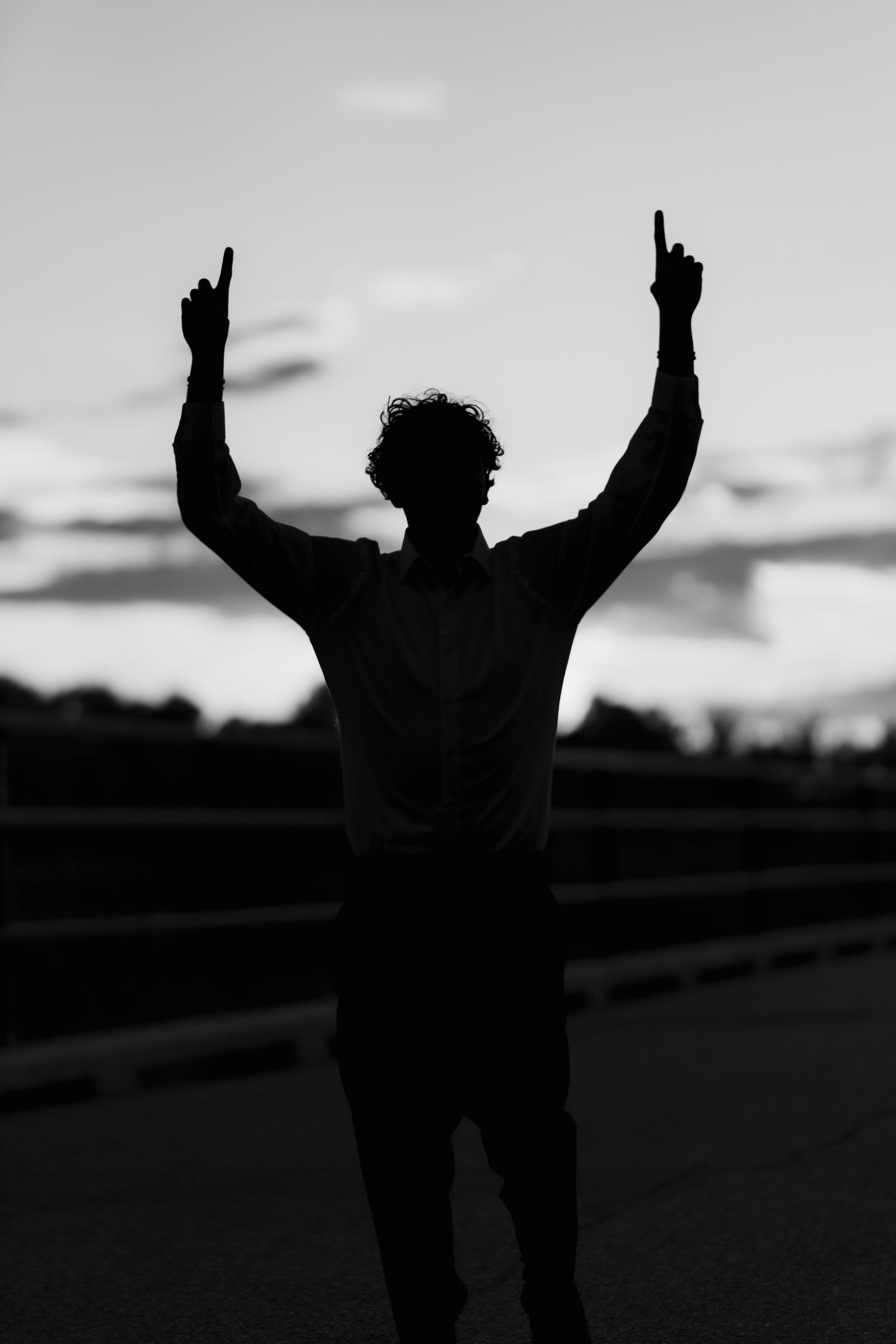 Silhouette of person raising both arms with index fingers pointed skyward against a dramatic dusk sky.