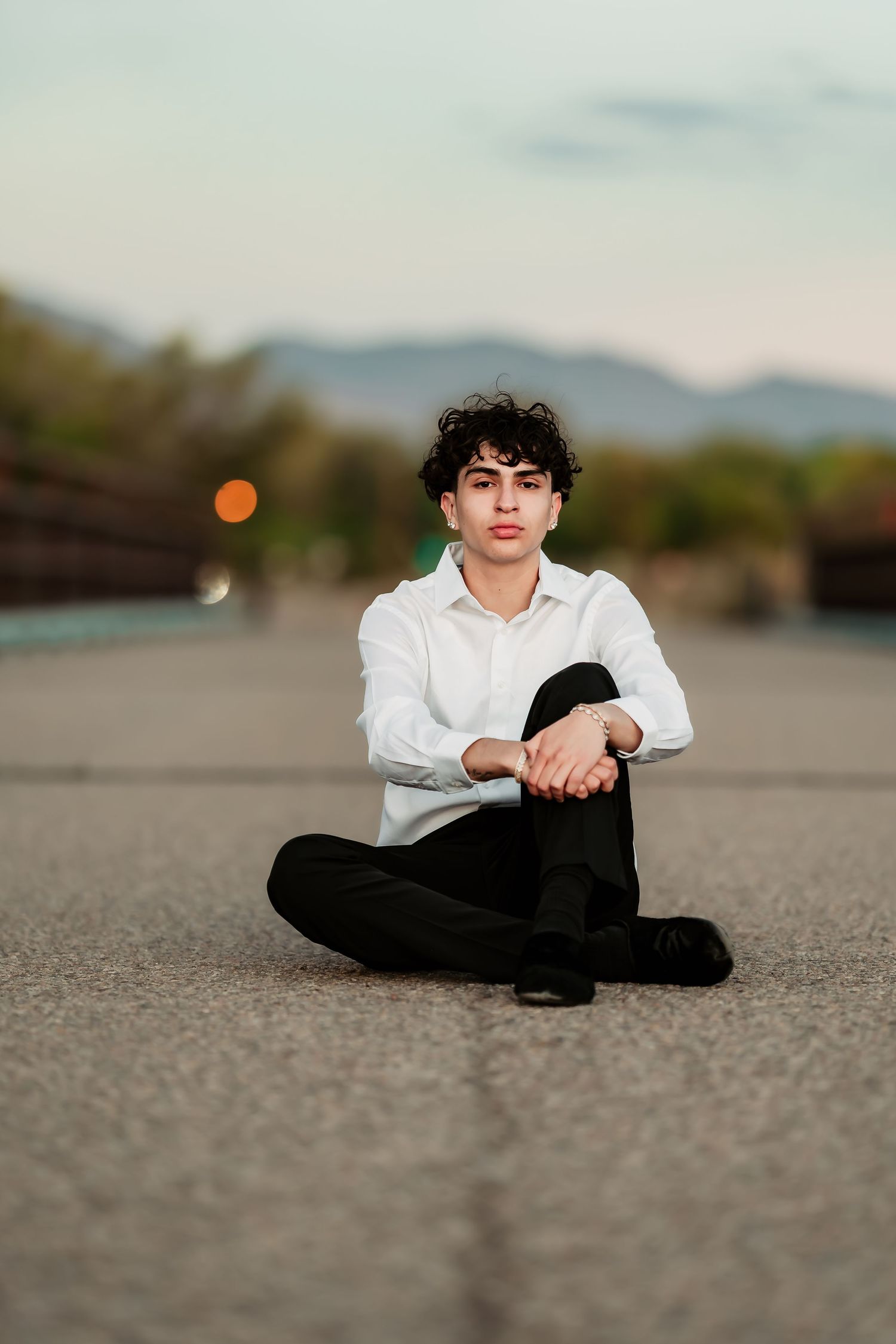 Portrait of a casual figure sitting cross-legged on the ground wearing a white shirt and black pants at sunset.