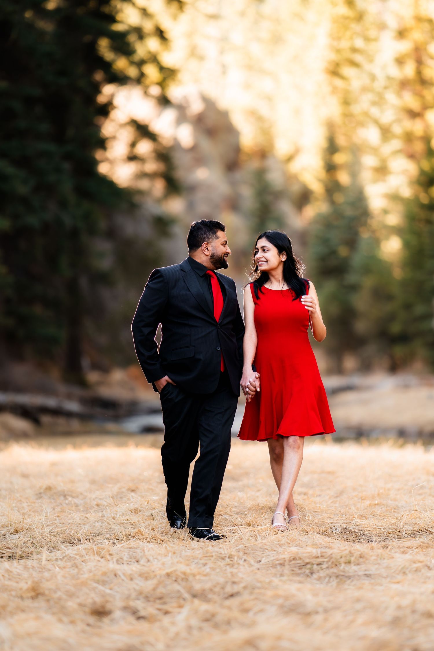 A couple in matching red and black formal attire walks together in a sunlit forest clearing.