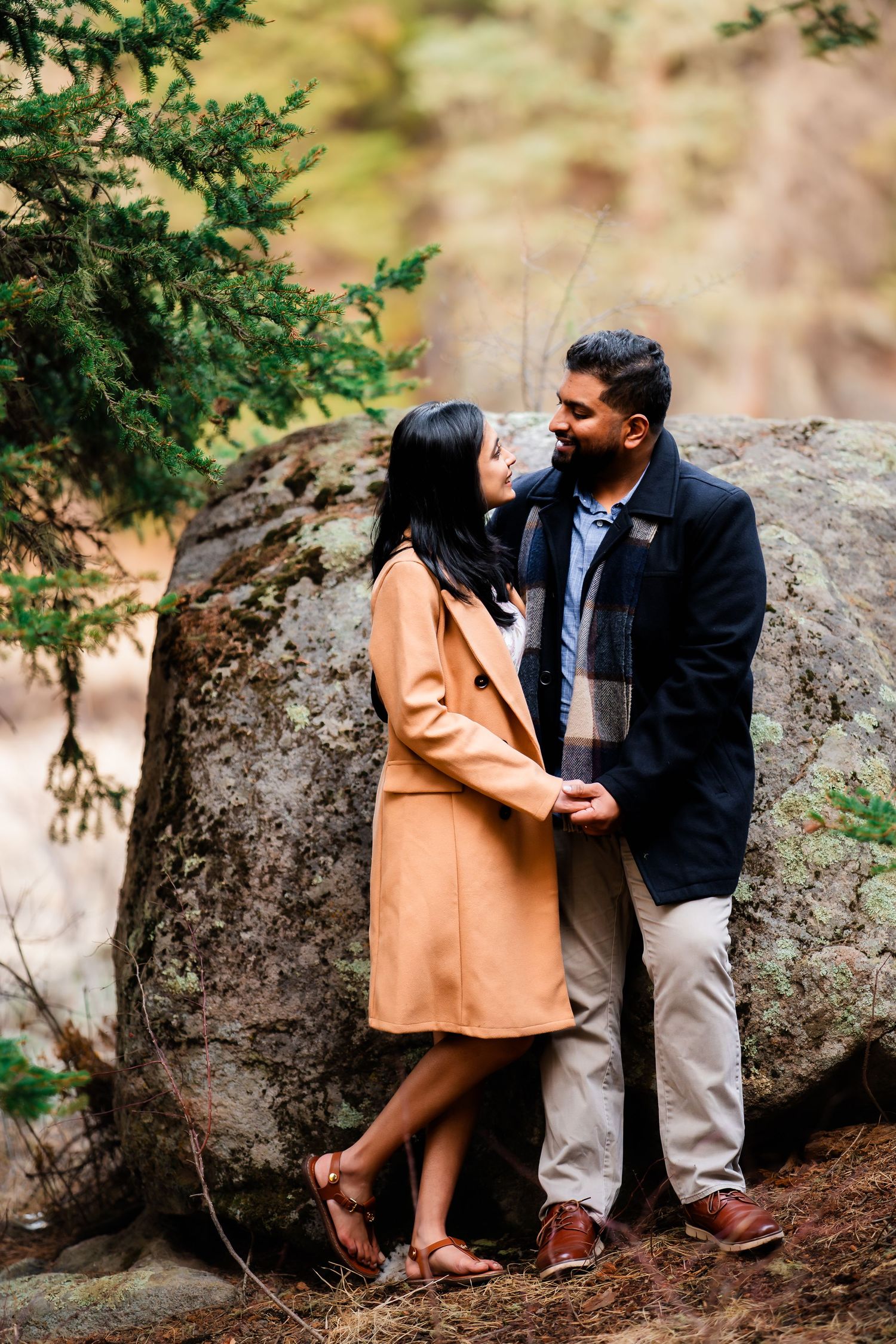 A romantic couple shares an intimate moment while standing by a large rock in a wooded area.