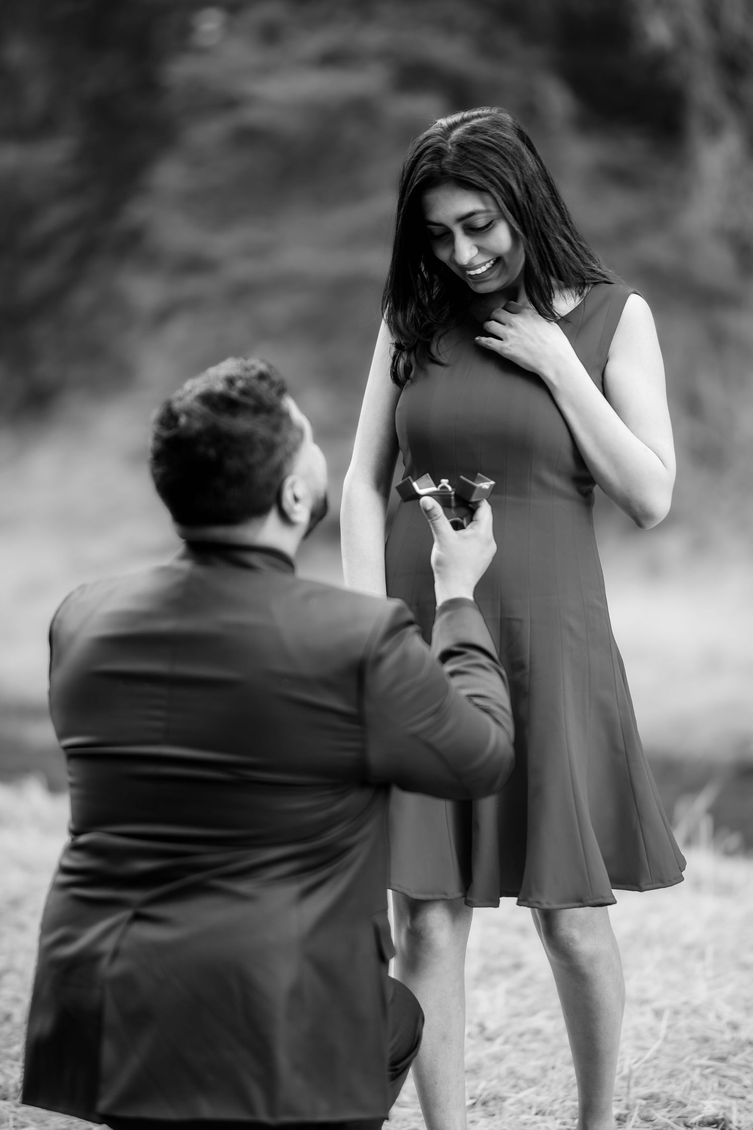 A tender marriage proposal moment captured in black and white as a man kneels before woman outdoors.