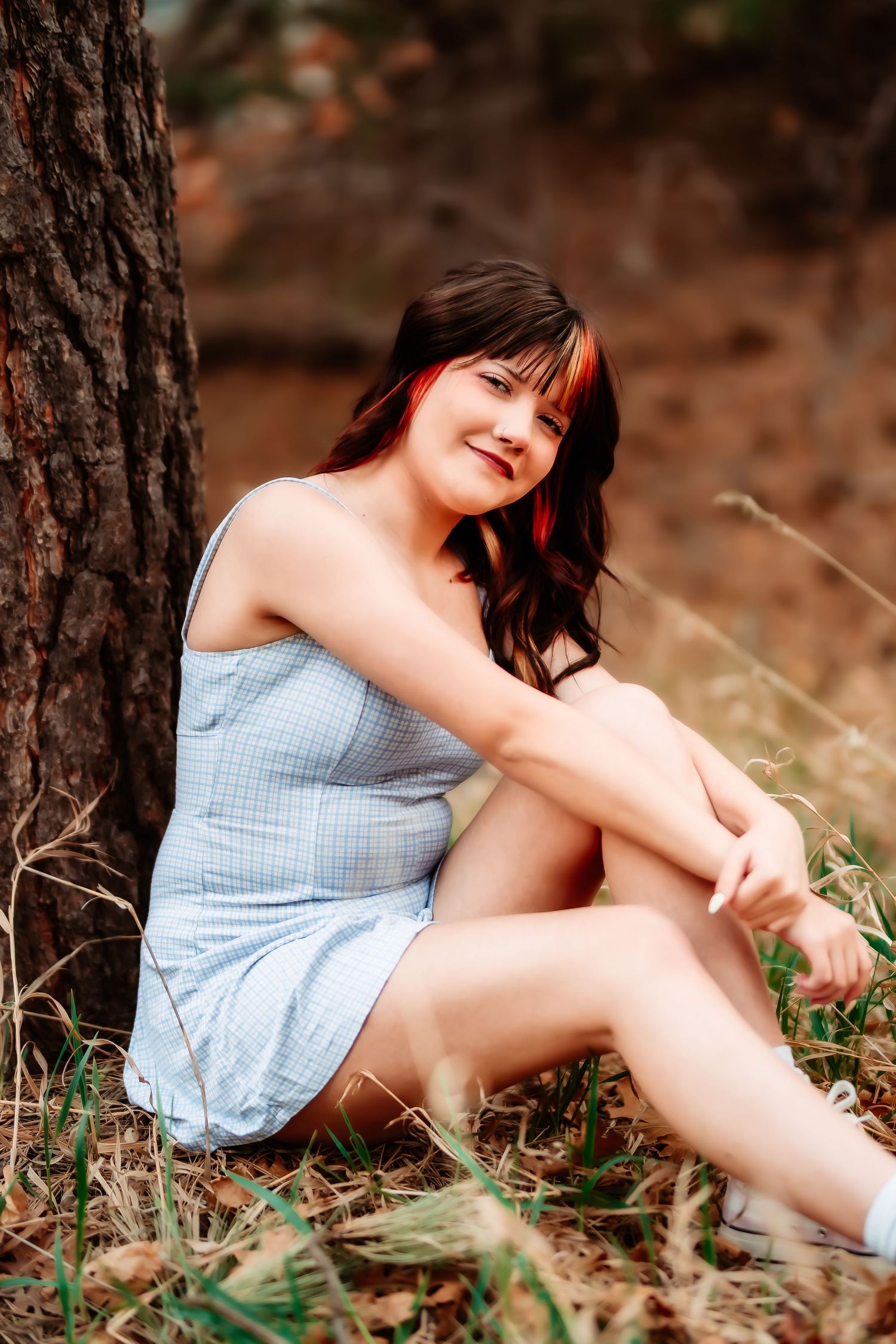 A person in a light blue dress sits against a tree trunk in a natural outdoor setting with warm lighting.