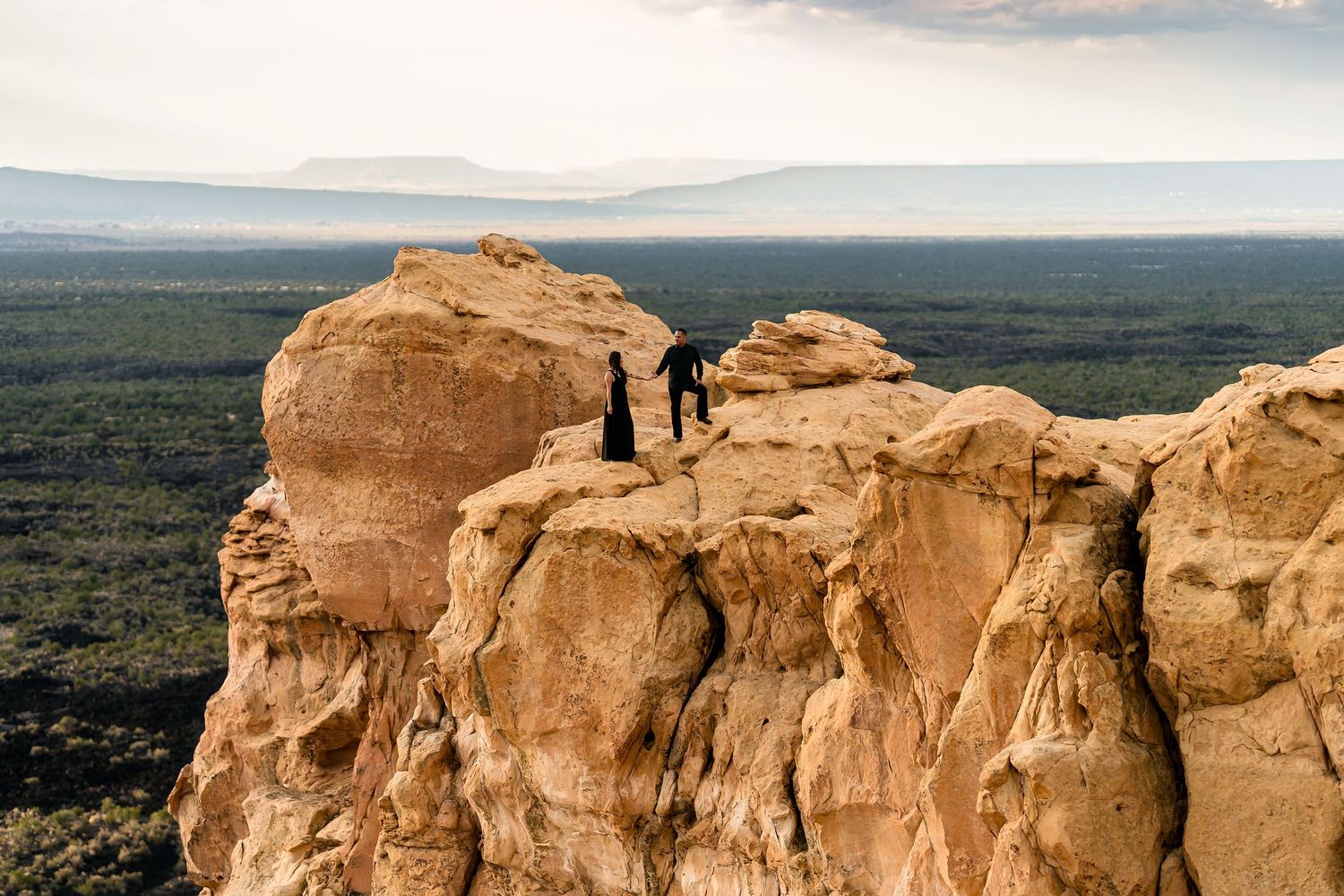 Two people standing on a dramatic sandstone cliff edge overlooking a vast desert landscape with distant mountains.