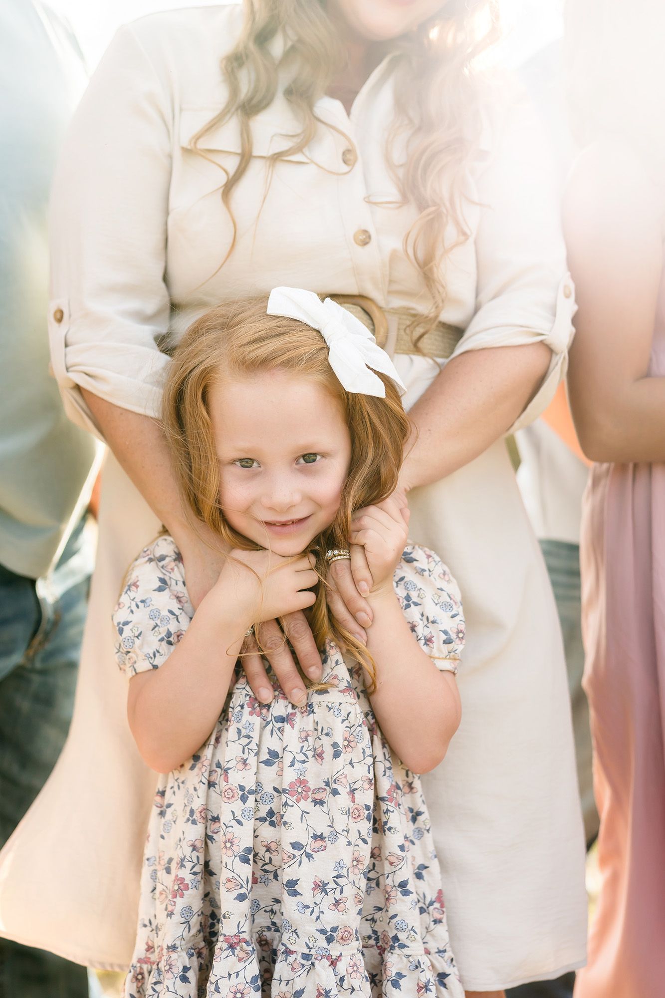 Young girl in floral dress with white bow smiles warmly while being embraced by mother in soft natural light.