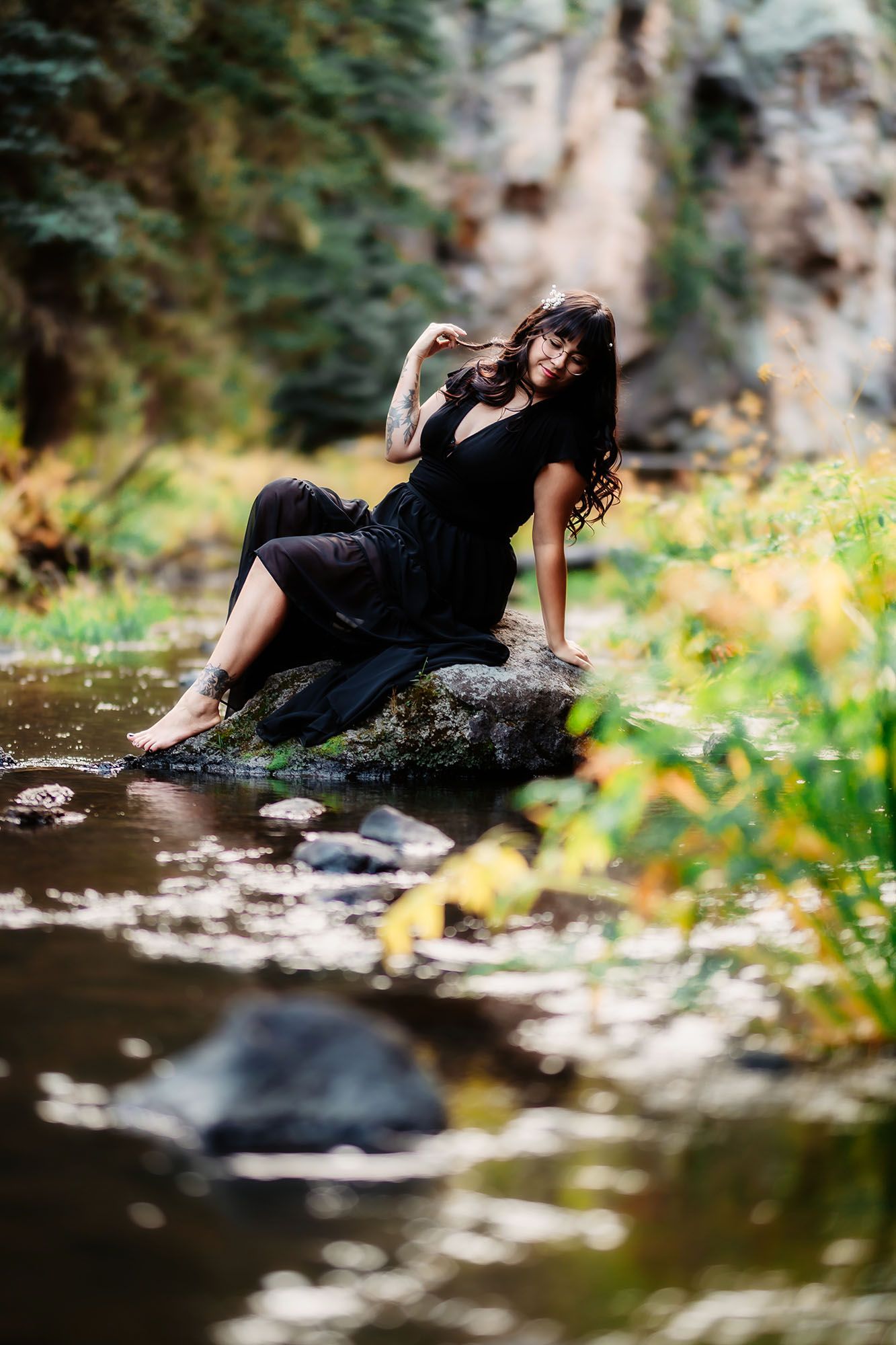 Tattooed woman in black dress sits barefoot on rocks beside a forest stream.