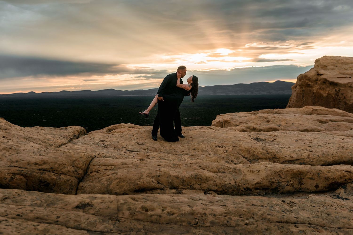 Couple sharing a dramatic dip kiss on sandstone rocks at sunset with mountain views behind them.