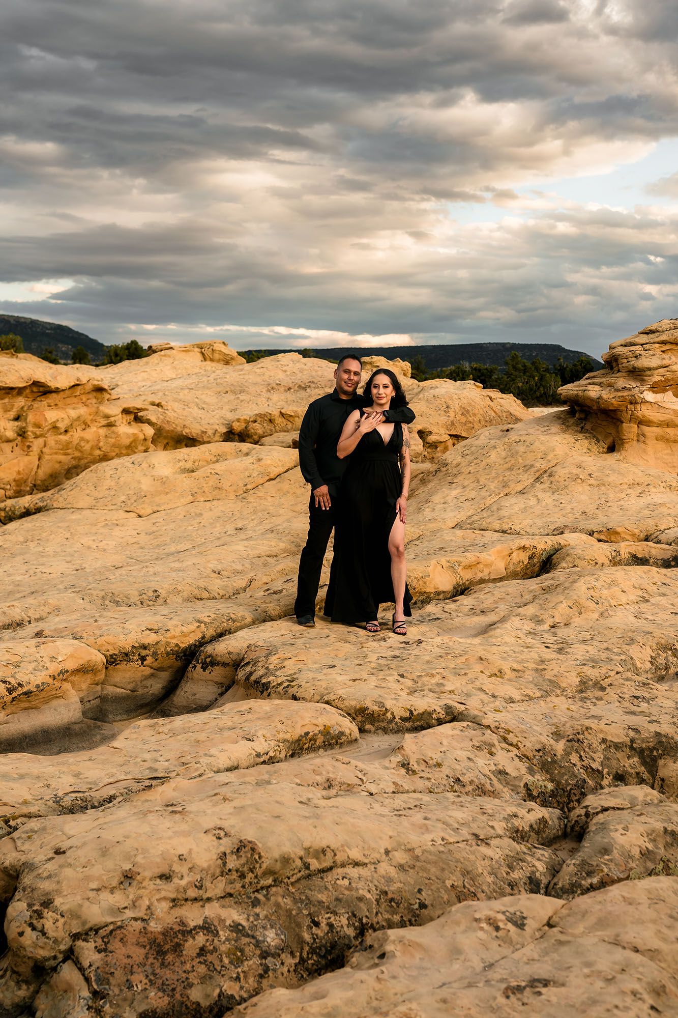 Couple posing together on rugged sandstone rocks wearing black outfits under a dramatic cloudy sunset sky.