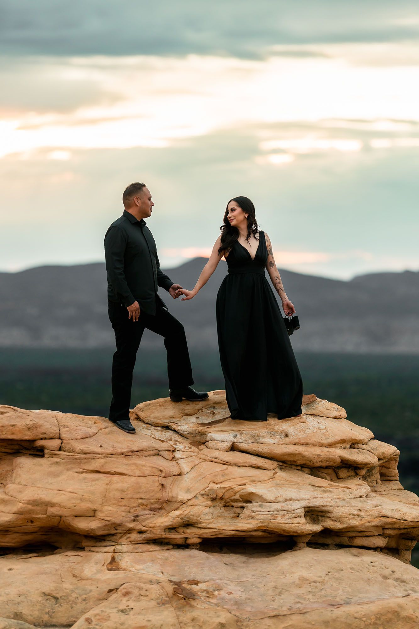 Couple in black formal attire holding hands on sandstone rock formation at sunset.