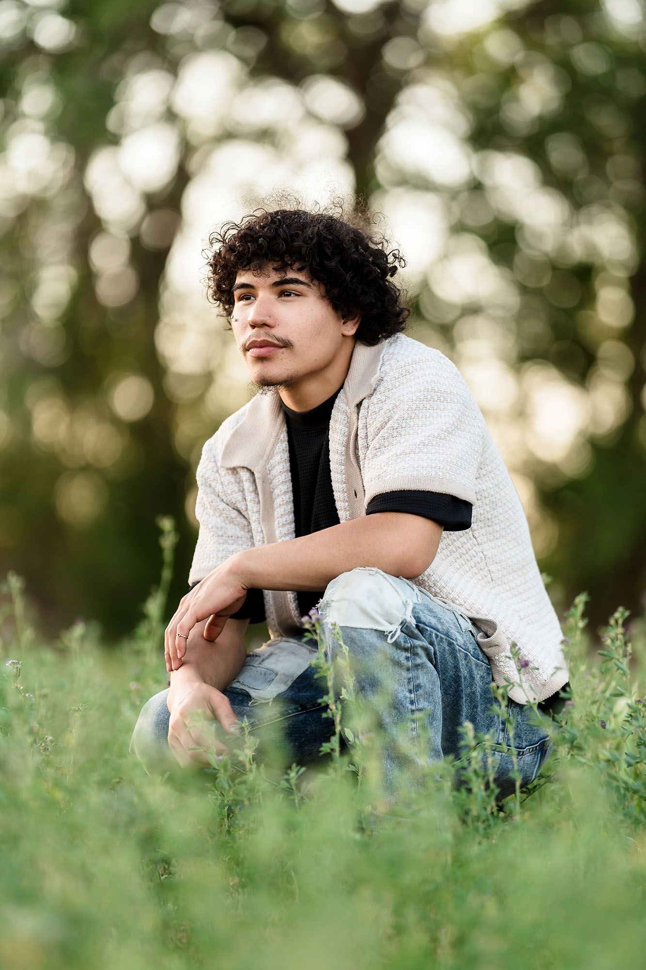 Young man with curly hair sitting in green grass outdoors, wearing jeans and a white top, looking thoughtfully to the side.