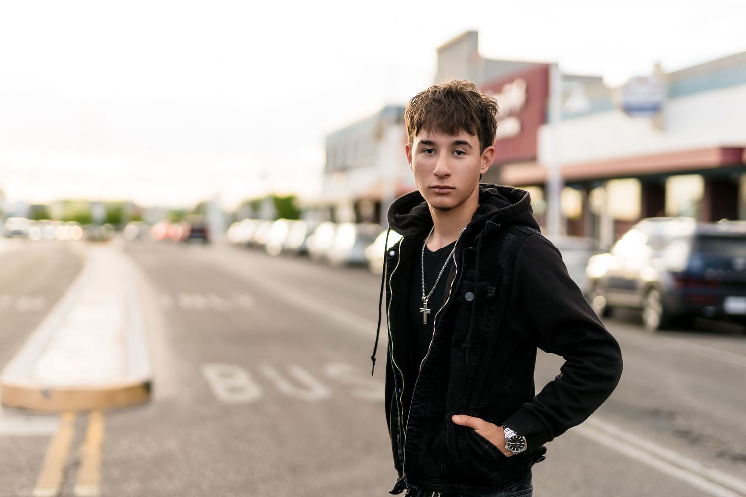 Young man in black hoodie posing on urban street with cars and storefronts in background.