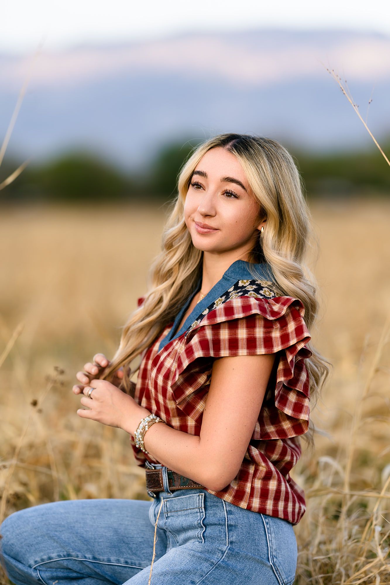 Blonde teenage girl in plaid top and jeans poses in golden wheat field at sunset.