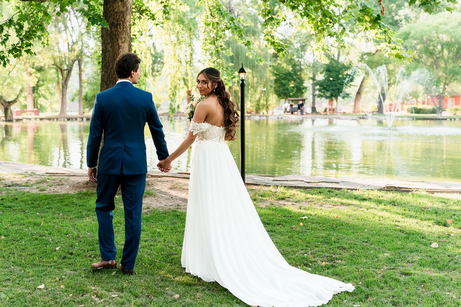 Bride and groom holding hands by a pond in a lush green park, wearing a white gown and navy suit.
