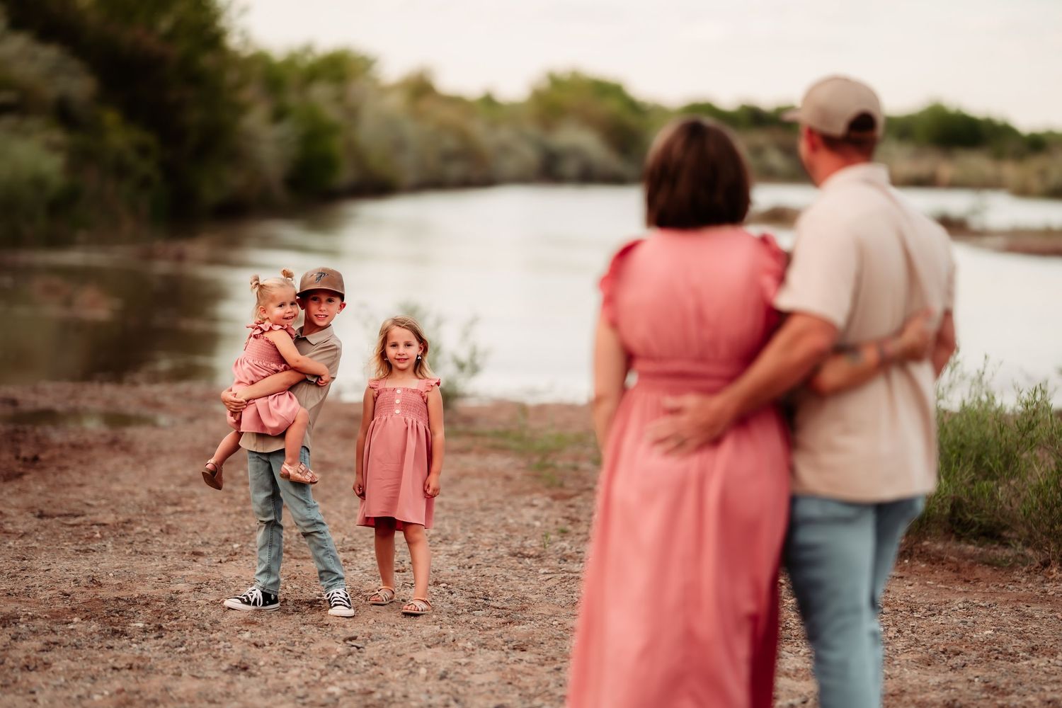 A family stands together on a riverbank at sunset wearing coordinating pink and casual outfits.