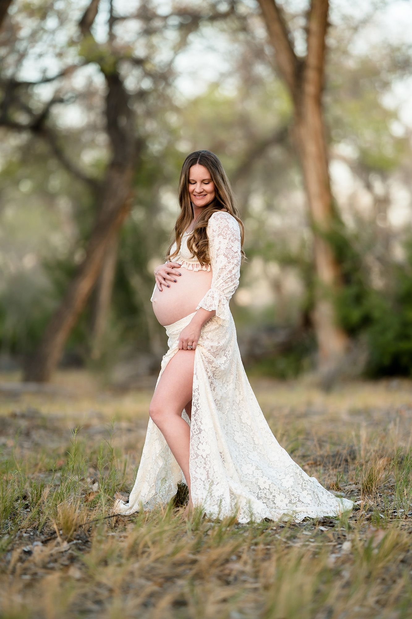 Pregnant woman in white lace maternity gown outdoors, cradling baby bump, smiling softly in a natural wooded setting.