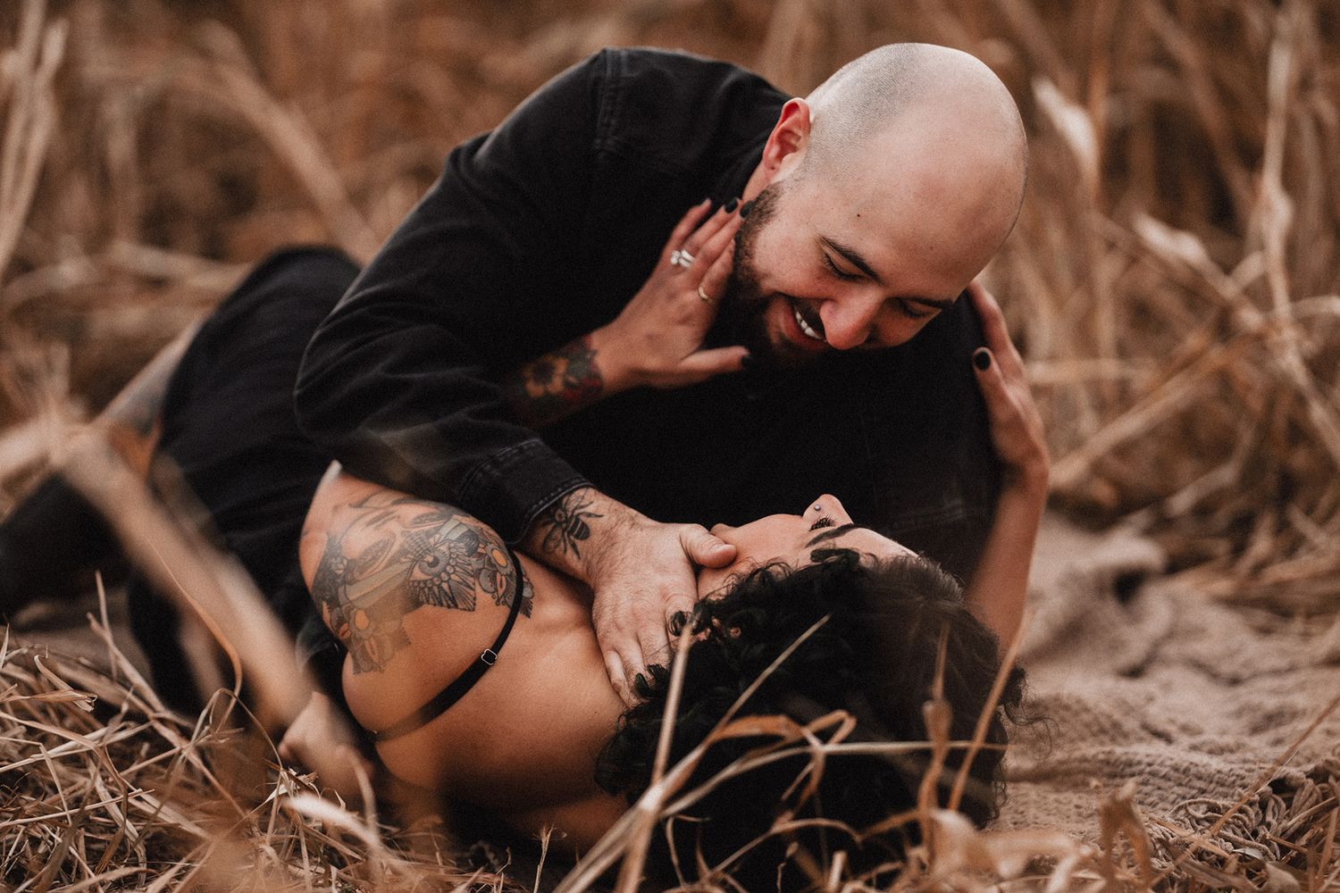 Tattooed bald man smiling and embracing a woman lying in golden dried grass field.