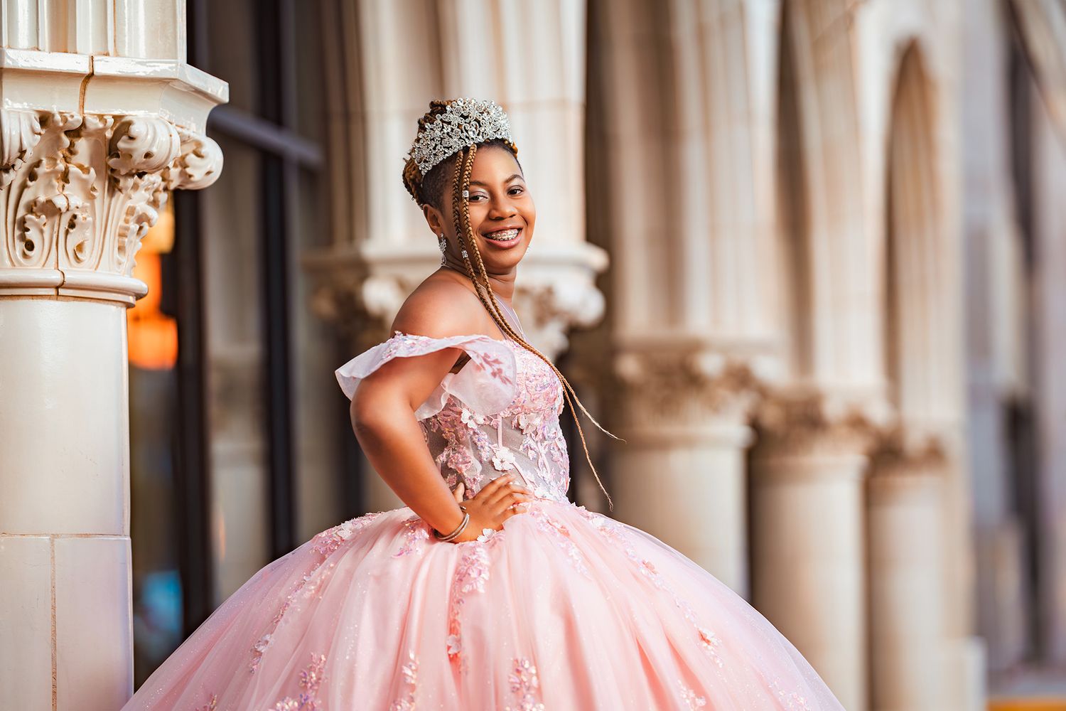 A person wearing a sparkling crown and pink princess ballgown poses gracefully between ornate stone columns.