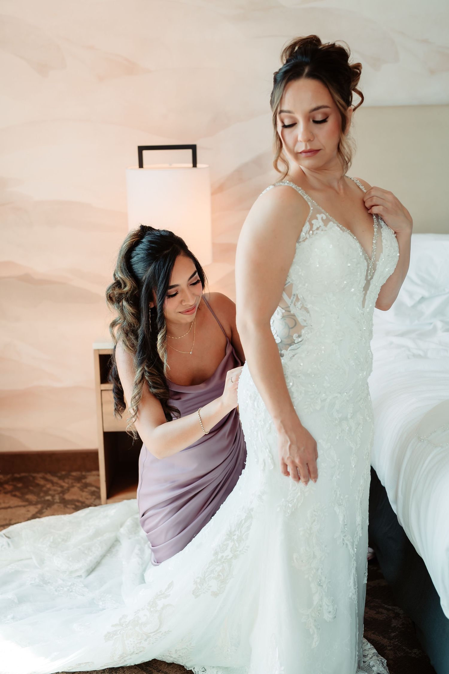 A bridesmaid in purple assists with fitting a detailed white lace wedding gown during wedding preparations.