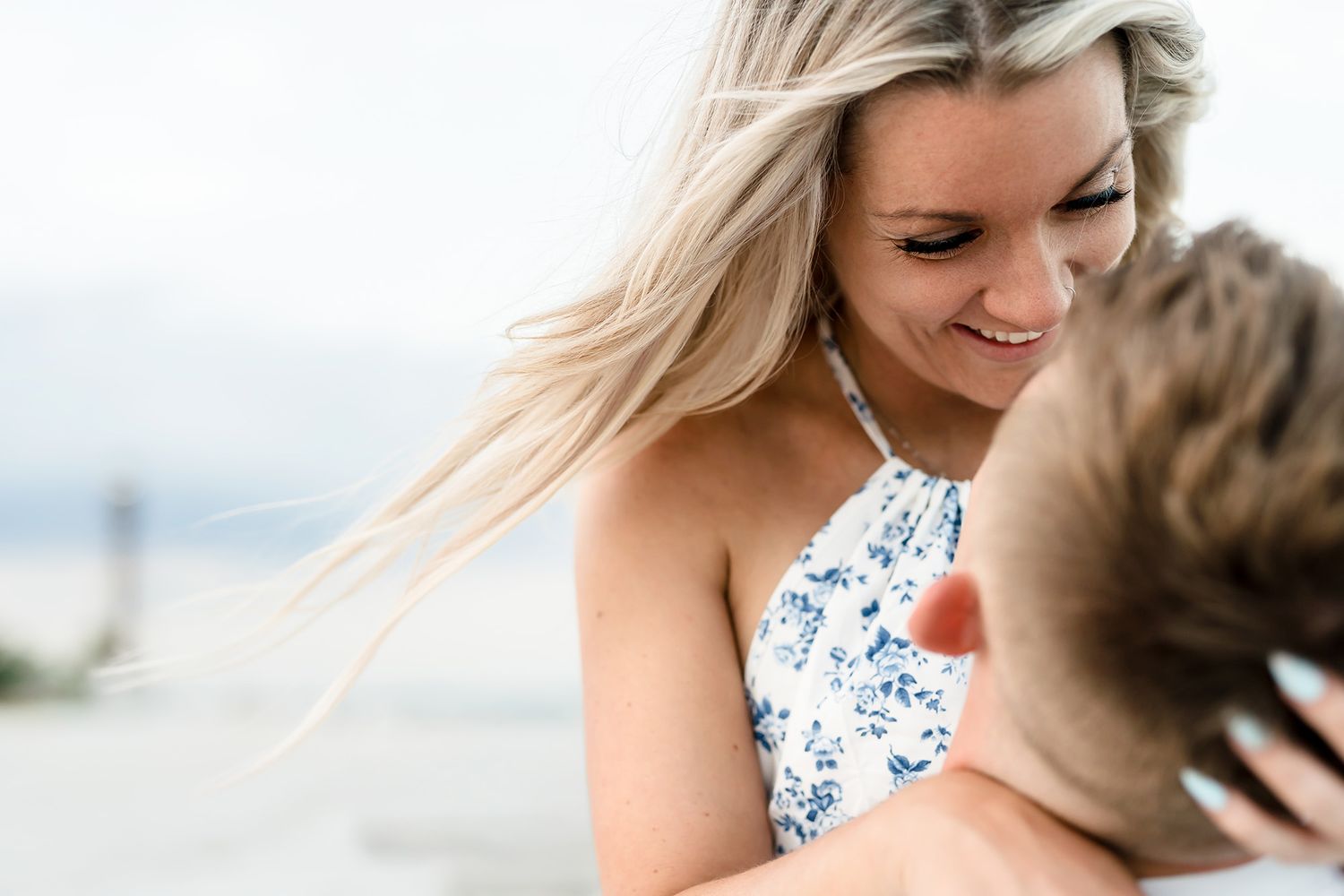 A smiling blonde woman in a floral halter top shares a romantic moment outdoors near the water.