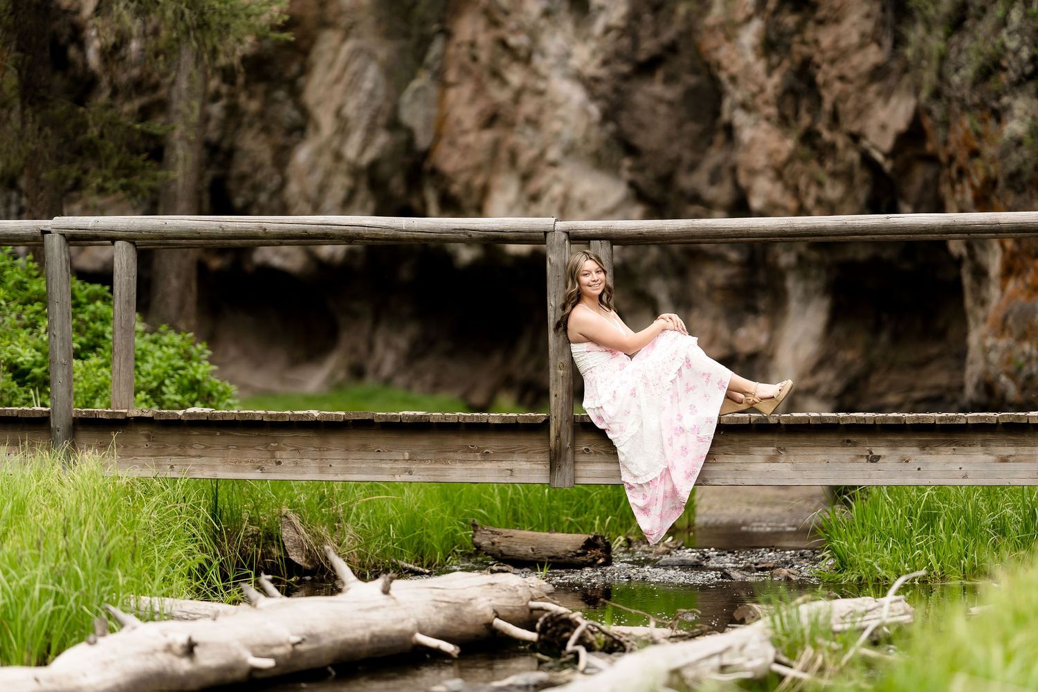 Woman in flowing pink dress posing on rustic wooden bridge surrounded by lush greenery and rocky cliffs.