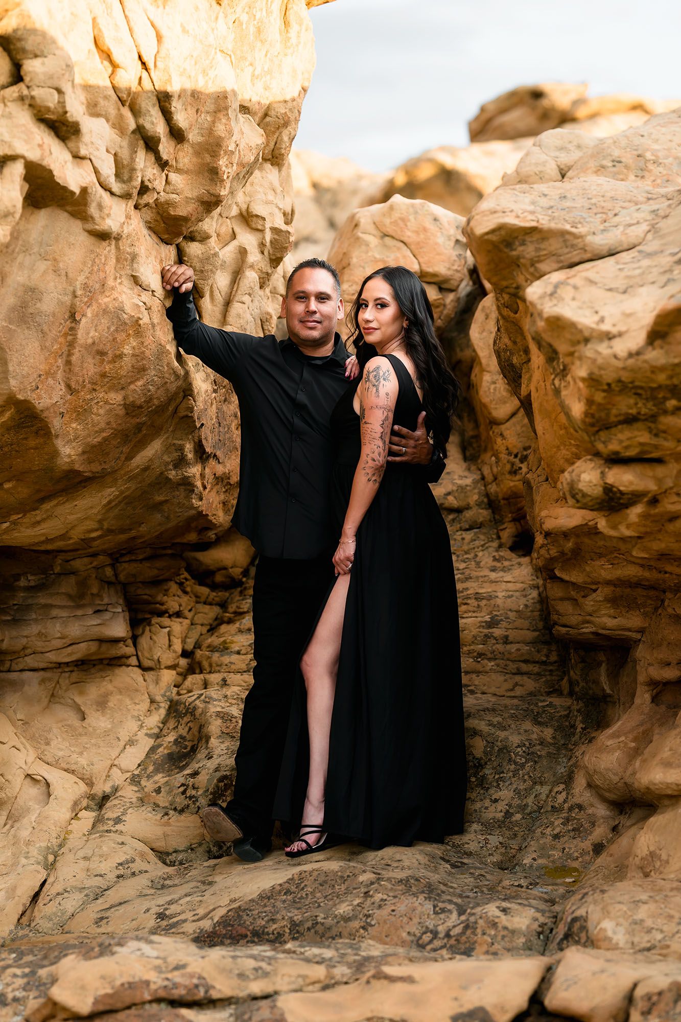 Couple in matching black outfits posing between golden sandstone rock formations in a romantic outdoor photo shoot.