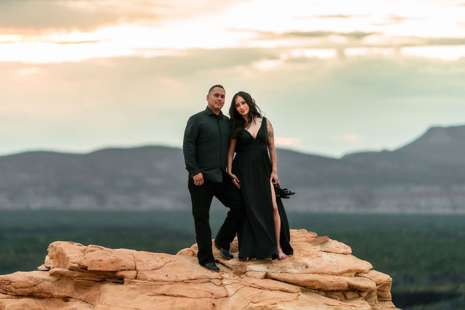 Couple in black outfits posing on red rock formation at sunset with mountain views in Sedona, Arizona.