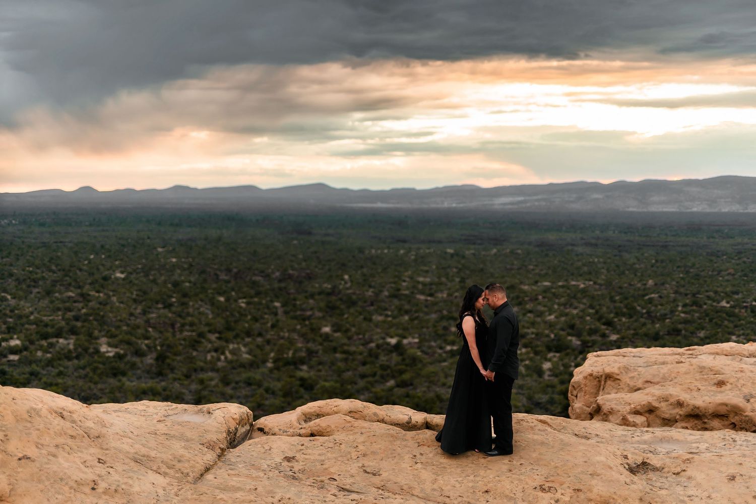Couple in black embracing on sandstone cliff overlooking vast desert valley at sunset.