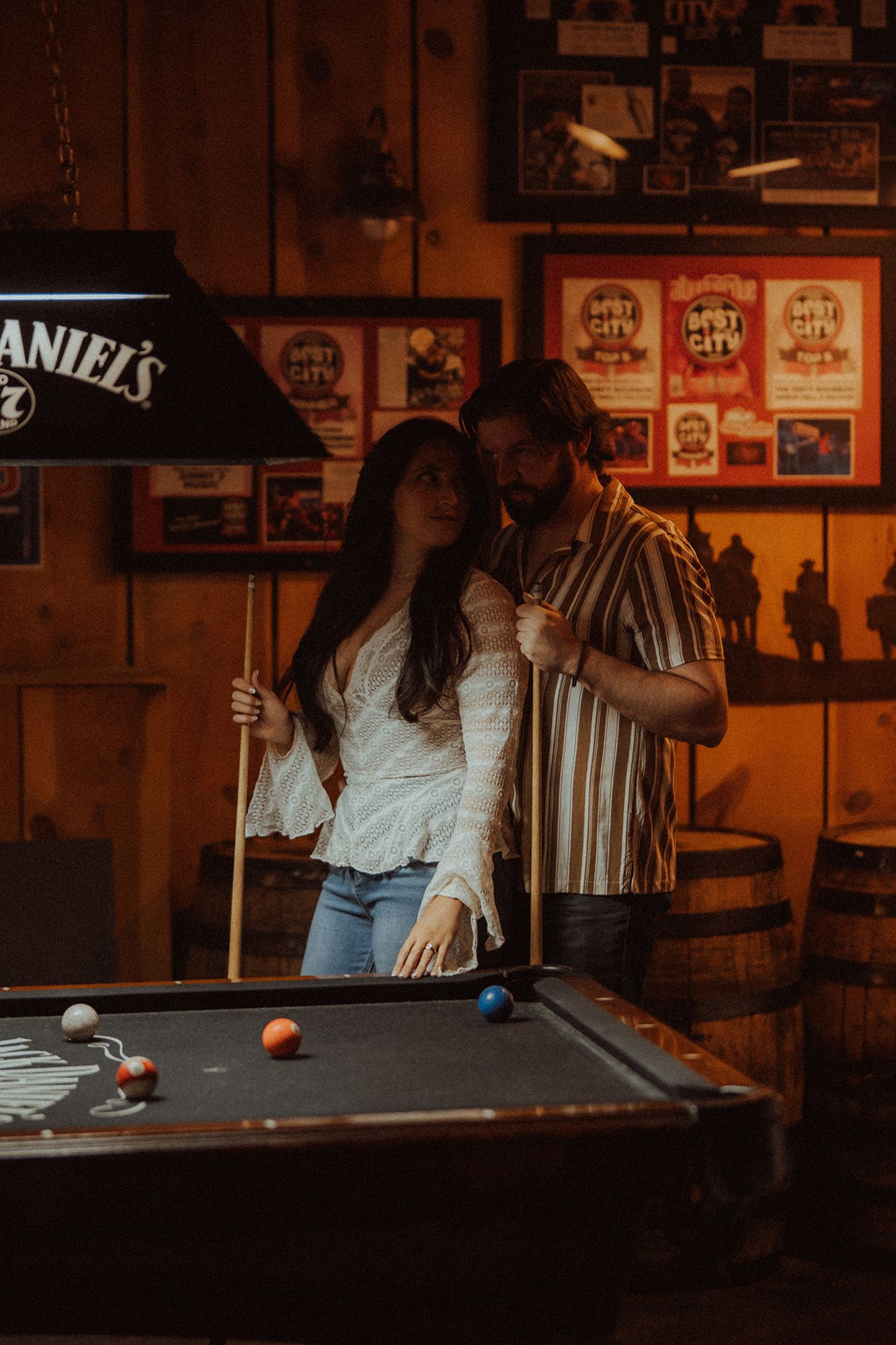 Couple playing pool in a rustic bar with Jack Daniel's lamp and warm lighting.