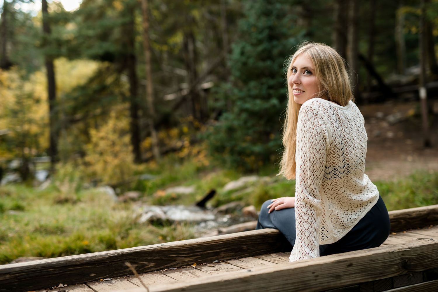 Blonde woman sitting on a wooden bridge smiling over her shoulder with autumn forest in background.