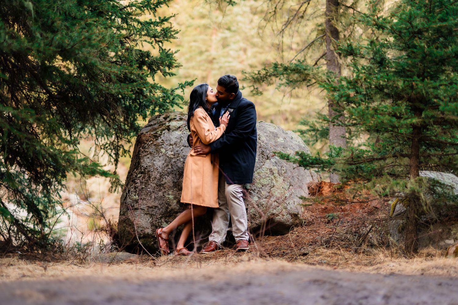 A loving couple shares an intimate moment near a large boulder in a forest setting.