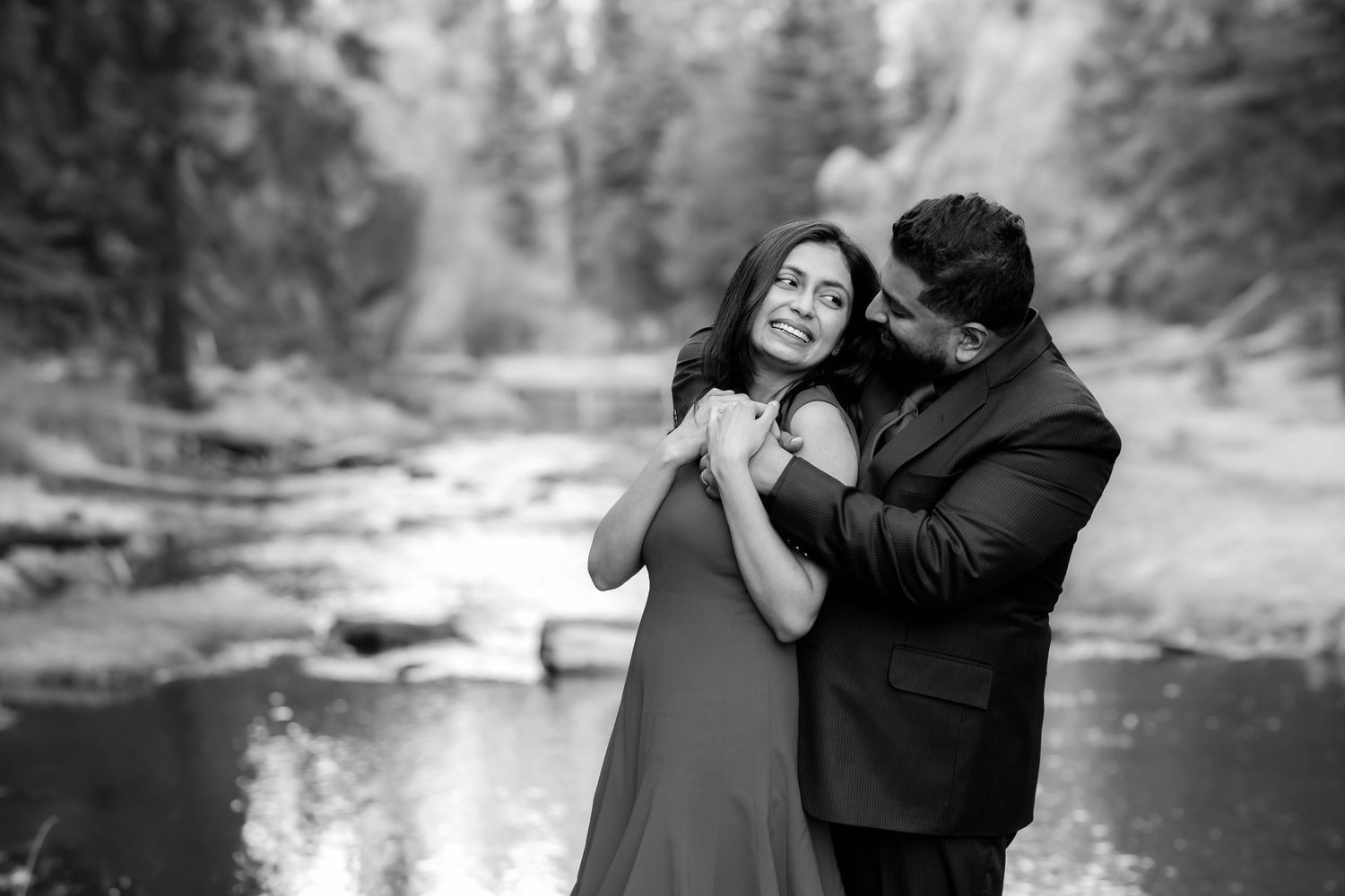 A couple shares a tender moment in black and white photography with a forest stream in the background.