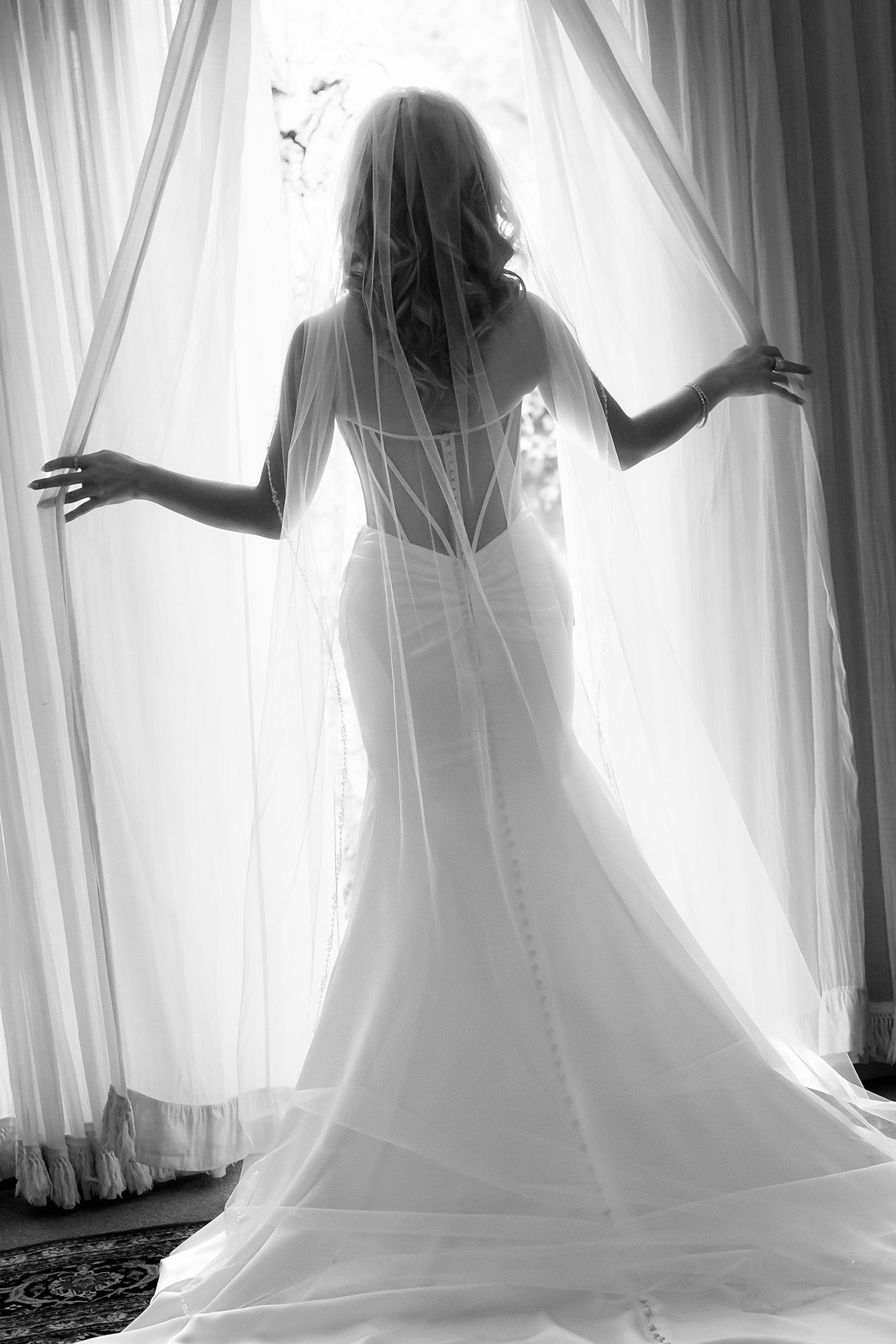 Black and white photo of a bride in a backless mermaid gown with veil, standing gracefully before a sunlit window.