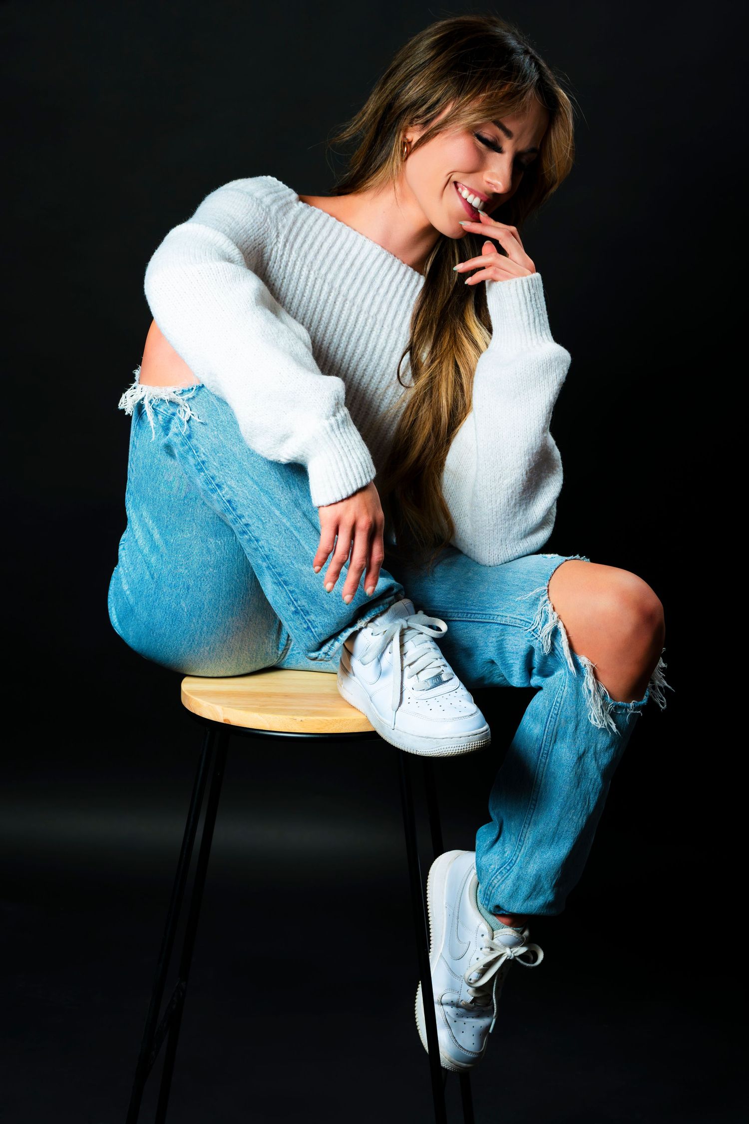 Person in white sweater and ripped jeans sits on stool wearing white sneakers against dark background in artistic poses.