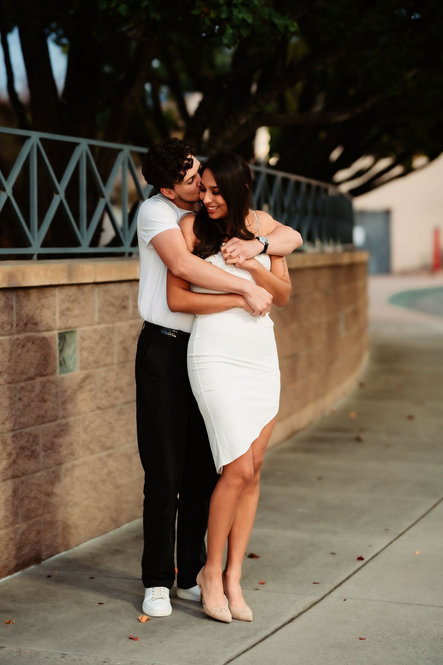 A couple in formal attire shares a romantic moment on a sidewalk against a stone wall.